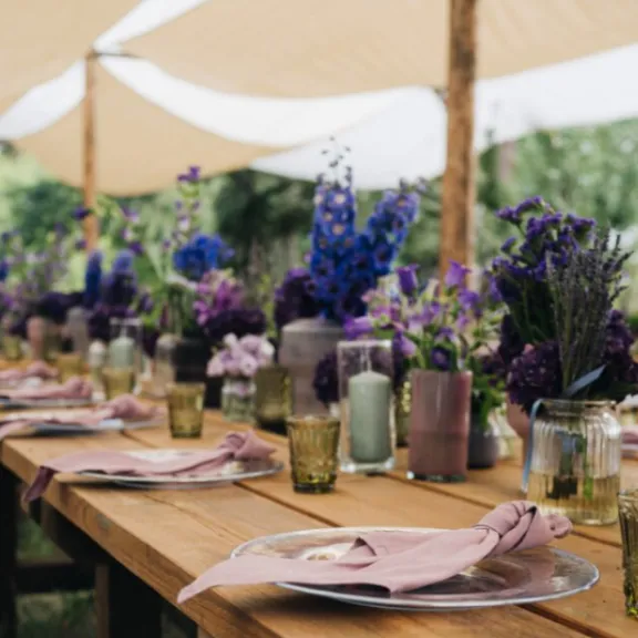 Lange Holztische mit lila Blumen und Gedecken im Freien, unter Sonnensegeln.