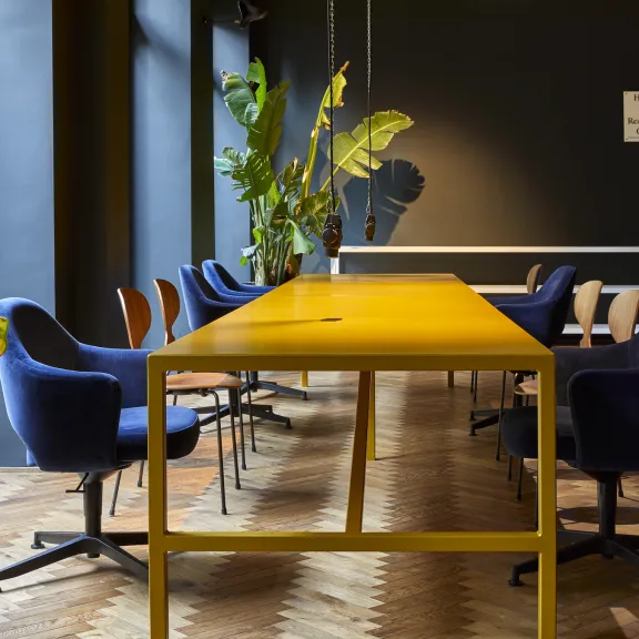 Co-Working Space mit gelbem Tisch, blauen Stühlen und großen Zimmerpflanzen.