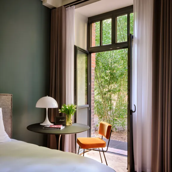 Helles Schlafzimmer mit offenem Fenster, Tisch, Stuhl und grünem Ausblick.