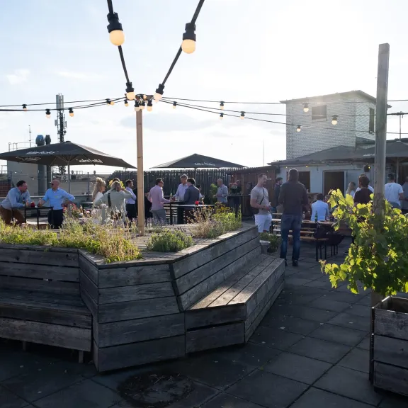 Dachterrasse mit Menschen, Holzbänken, Pflanzen und Lichterketten bei Sonnenschein.