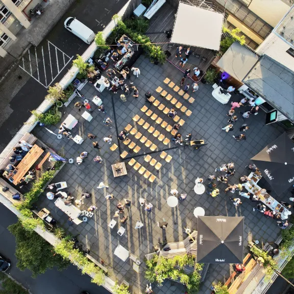 Dachterrasse mit Stühlen, Menschen und Tischen, umgeben von Gebäuden und Bäumen