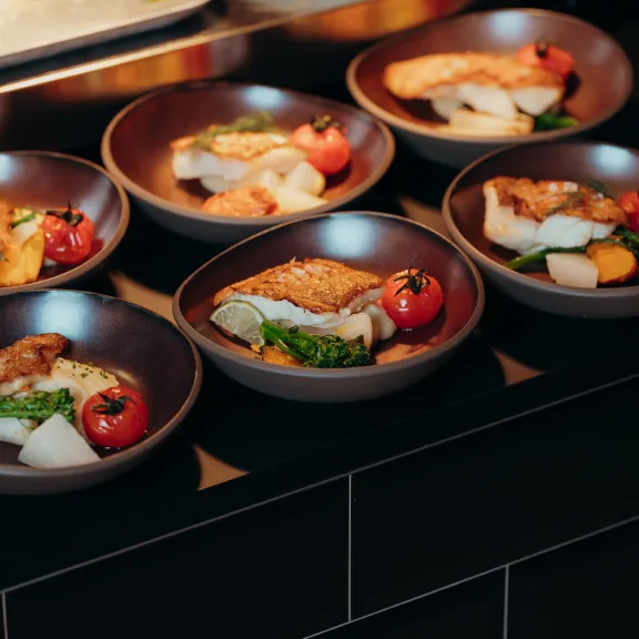 Sechs Teller mit Fischfilet, Gemüse und Kirschtomaten auf schwarzer Theke.