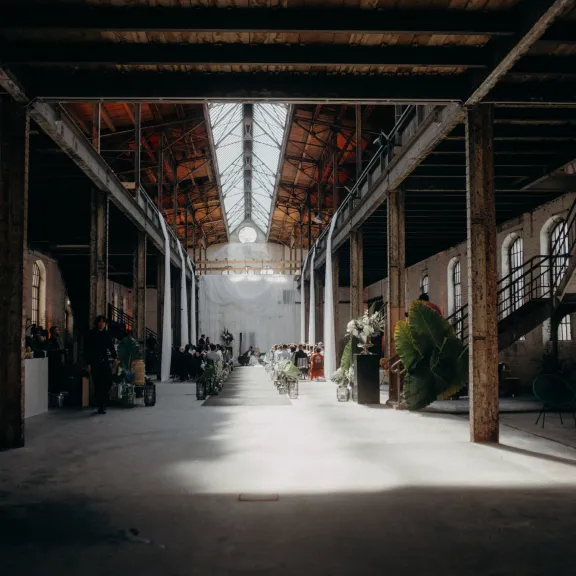 Lichtdurchflutete Industriehalle mit Pflanzen, Möbeln und einer Treppe rechts.