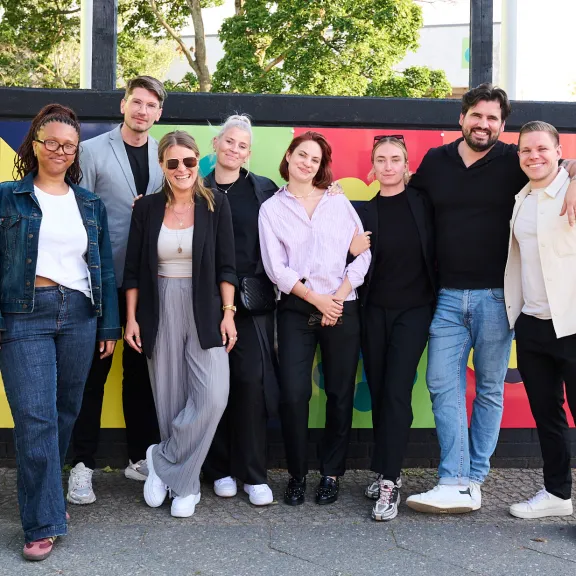 Acht Personen stehen lächelnd vor einer bunten Wand im Freien.