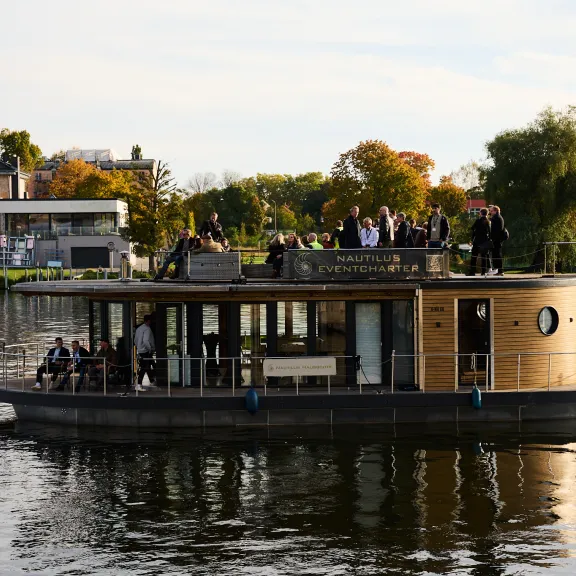 Hausboot mit Menschen fährt auf einem Fluss, Bäume und Häuser im Hintergrund