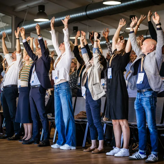 Viele Menschen stehen von ihren Stühlen auf und strecken ihre Arme in die Höhe.