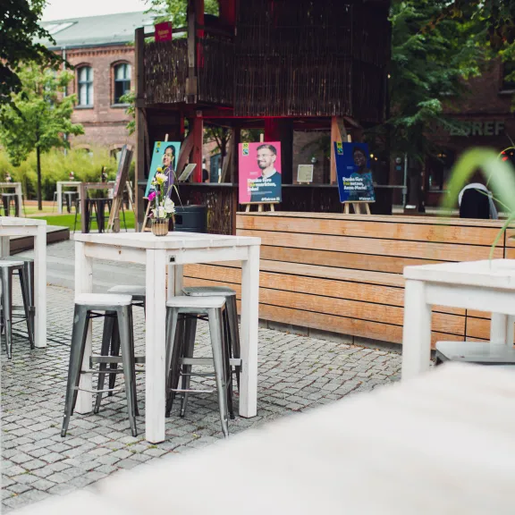 Weiße Tische und Stühle auf einer Terrasse, im Hintergrund Bäume und ein Holzgebäude.