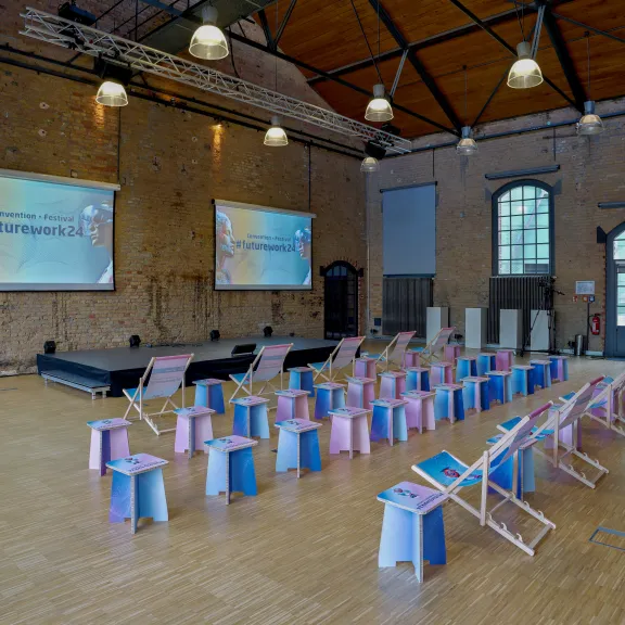 Veranstaltungssaal mit bunten Stühlen, Bühne und zwei großen Leinwänden