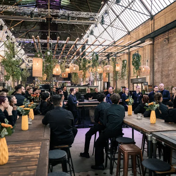 Große Gruppe Menschen sitzt in einer Halle an langen Holztischen mit gelben Vasen.