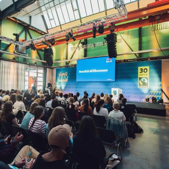 Viele Menschen sitzen in einem modernen Saal und schauen auf eine große Präsentationsleinwand.