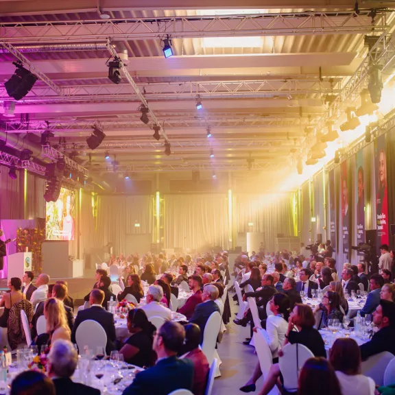 Großer Saal mit festlich gekleideten Menschen bei einer Gala, bunt beleuchtet.