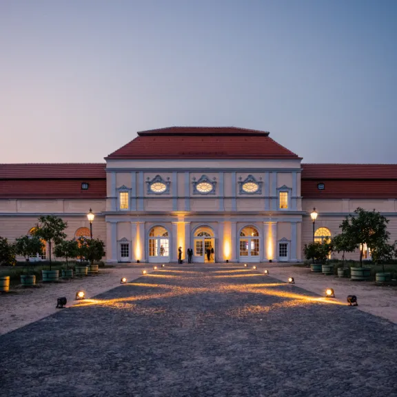 Beleuchtetes barockes Gebäude mit rotem Dach und symmetrischem Vorplatz bei Dämmerung.