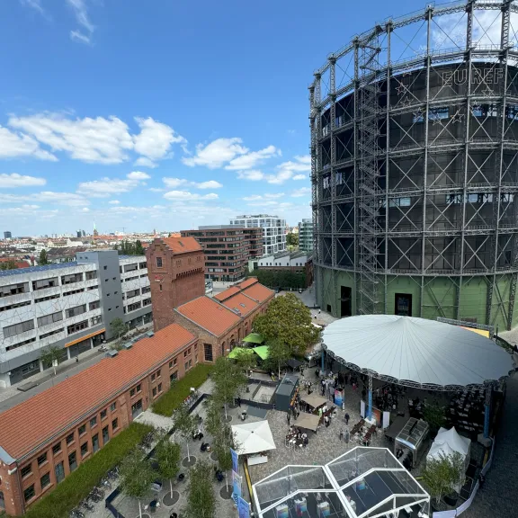 Großer runder Gasometer, rote Backsteingebäude, Menschen im Hof, blauer Himmel mit Wolken.