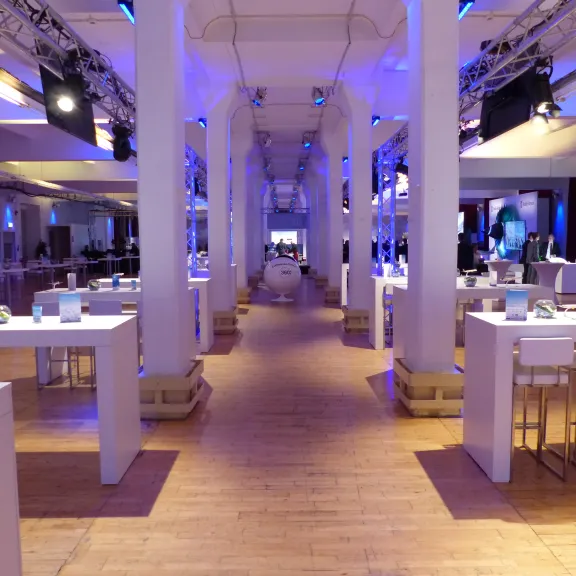 Großer, modern beleuchteter Ausstellungsraum mit weißen Tischen und Bildschirmen.