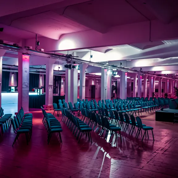 Reihe leerer Stühle in beleuchtetem Veranstaltungssaal mit Bühne und buntem Licht.