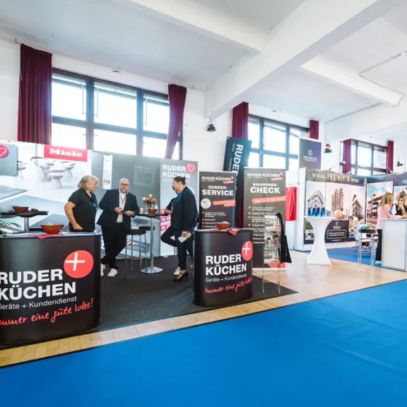 Messehalle mit mehreren Ständen, Menschen im Gespräch, blauer Teppichboden.
