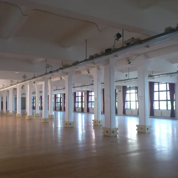 Heller, leerer Saal mit Holzboden, weißen Säulen und großen Fenstern