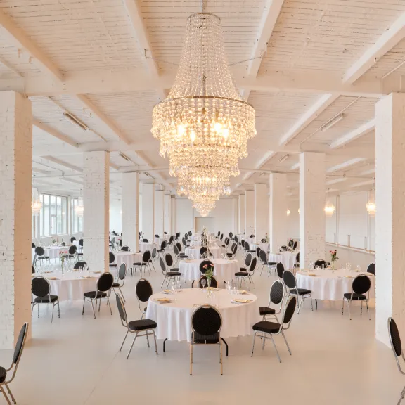 Großer, heller Festsaal mit runden Tischen, schwarzen Stühlen und Kronleuchtern.