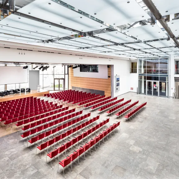 Großer, heller Hörsaal mit vielen roten Stuhlreihen und großer Fensterfront.