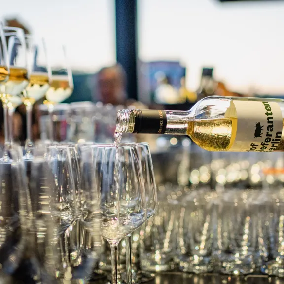 Weißwein wird in mehrere Gläser eingeschenkt, festliche Stimmung im Hintergrund.