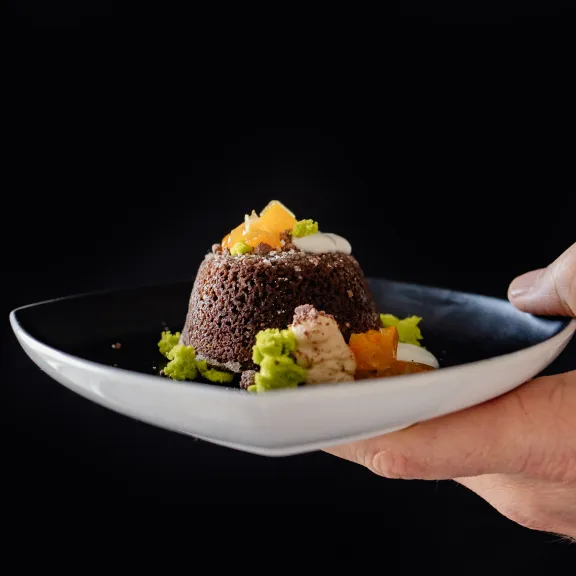 Hand hält einen Teller mit Schokoladendessert und bunten Obststücken vor schwarzem Hintergrund.