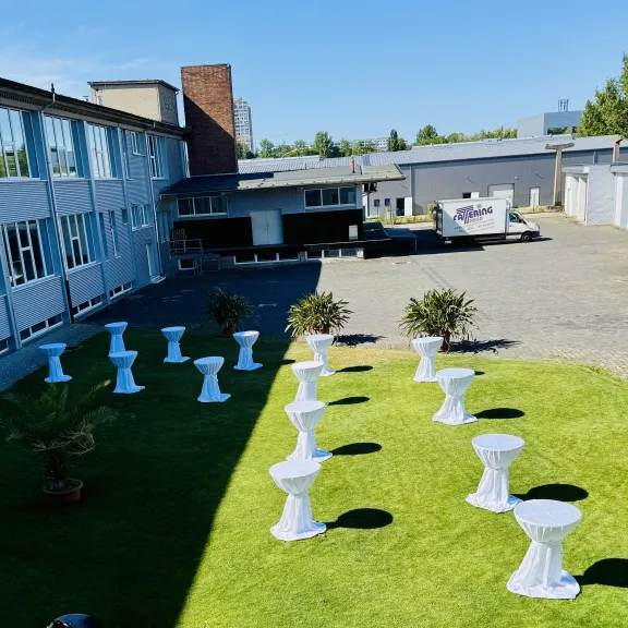 Stehtische mit weißen Hussen auf grünem Rasen neben modernen Gebäuden bei Sonnenschein.