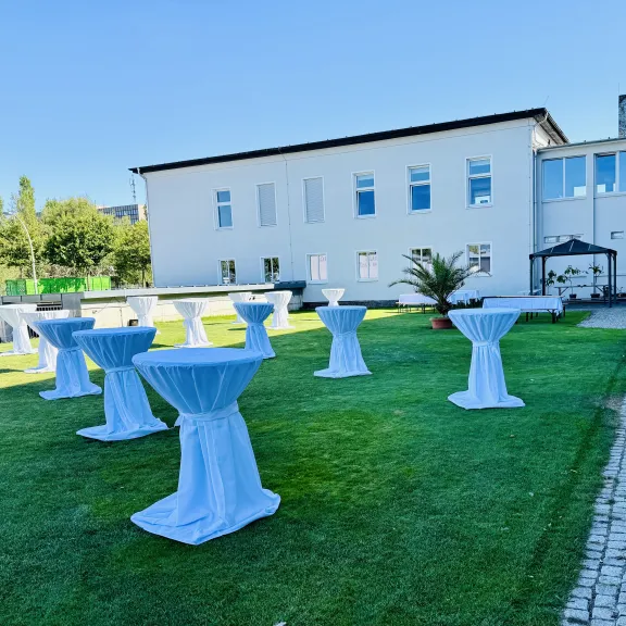 Stehtische mit weißen Hussen auf grünem Rasen vor einem modernen Gebäude bei Sonnenschein.