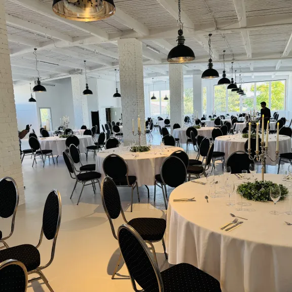 Heller, moderner Saal mit runden, festlich gedeckten Tischen und schwarzen Stühlen.