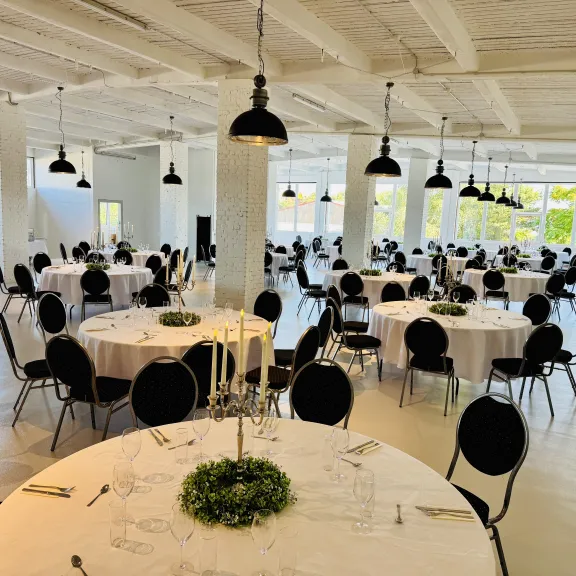 Heller Festsaal mit runden, festlich gedeckten Tischen und schwarzen Stühlen.