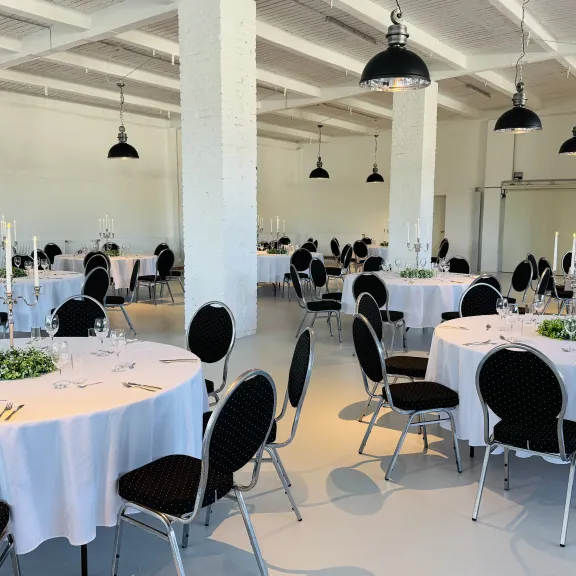 Runder Saal mit weißen, festlich gedeckten Tischen und schwarzen Stühlen.