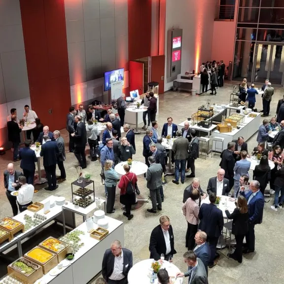 Menschen unterhalten sich an Stehtischen bei einer Konferenz mit Buffet im Hintergrund.