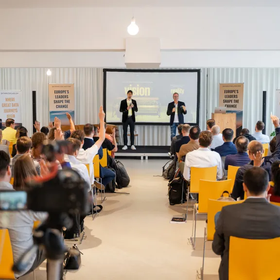 Menschen heben Hände bei einer Präsentation, zwei Redner auf Bühne, gelbe Stühle.