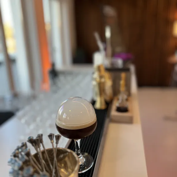 Cocktailglas mit Schaumkrone auf Bartheke, im Hintergrund Gläser und Barutensilien.