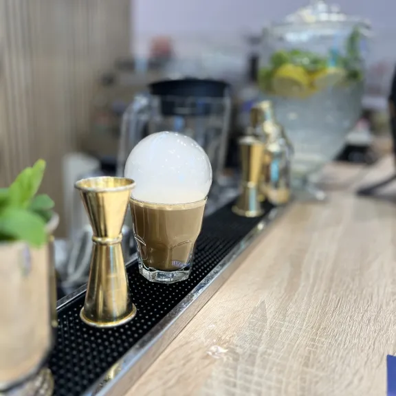 Latte mit Milchschaum auf Theke, daneben Messbecher und Glasgefäß mit Zitronenwasser.