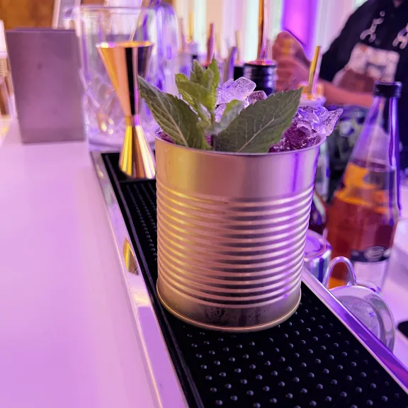 Cocktail in goldener Dose mit Eis und Minze auf einer Bar, lila Beleuchtung.