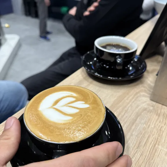 Cappuccino mit Latte Art und schwarze Tasse Kaffee auf Holztisch, Personen im Hintergrund.