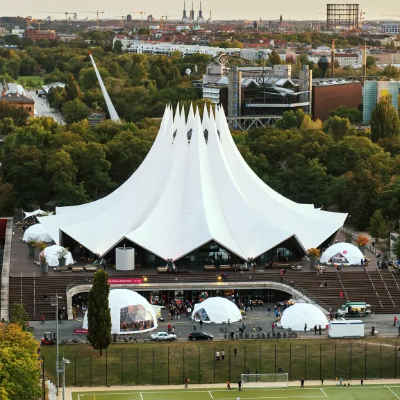 Weißes Zeltgebäude im Park, zahlreiche Besucher davor, Stadt im Hintergrund