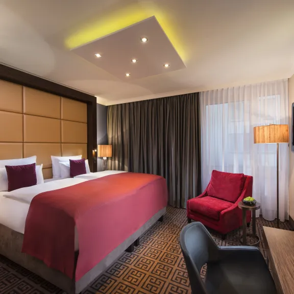 Modernes Hotelzimmer mit Doppelbett, rotem Sessel, Schreibtisch und warmem Licht.