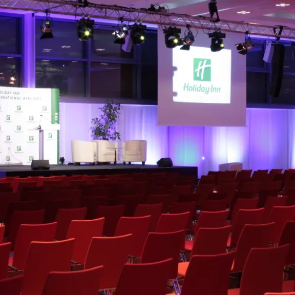 Leerer Konferenzraum mit roten Stühlen und beleuchteter Bühne, Holiday Inn Logo auf Leinwand.