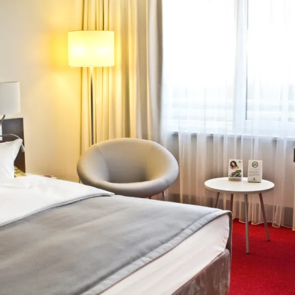 Helles Hotelzimmer mit Doppelbett, Sessel, Stehlampe und Schreibtisch auf rotem Teppich.