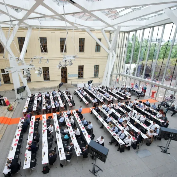 Großer Konferenzraum mit vielen Teilnehmern an langen Tischen und Glasdach.