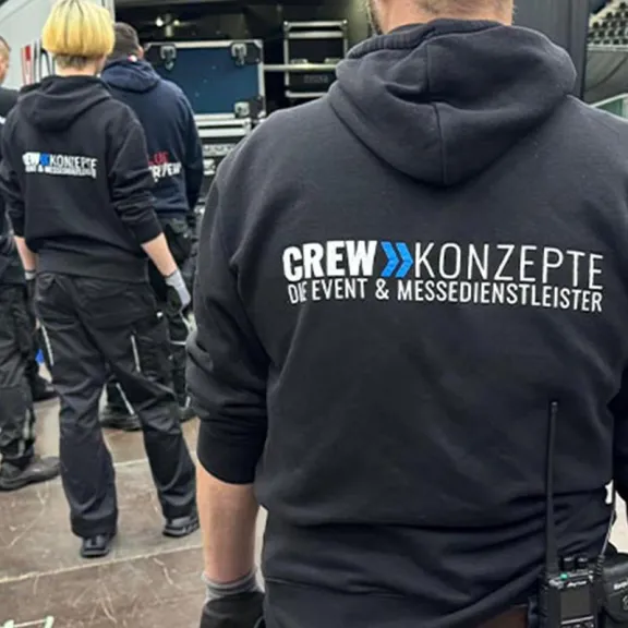 Zwei Crewmitglieder in schwarzen Hoodies mit Aufschrift, arbeiten an einer Veranstaltung.
