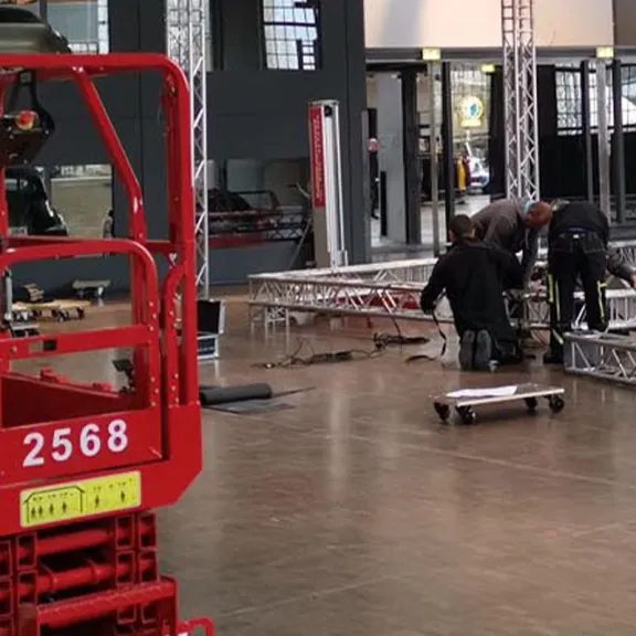 Arbeiter bauen eine Bühne in einer Halle auf, vorne steht eine rote Hebebühne.