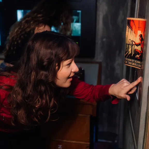 Frau mit braunen Haaren zeigt lächelnd auf ein Plakat an einer Wand, Kontrollraum im Hintergrund.