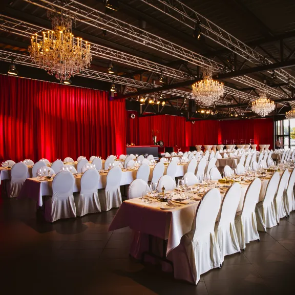 Eleganter Bankettsaal mit runden Tischen, weißen Stühlen und roten Vorhängen.
