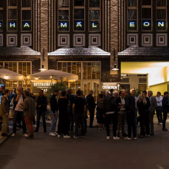 Menschenmenge vor einem beleuchteten Gebäude mit Schriftzug „Chamäleon“ am Abend.