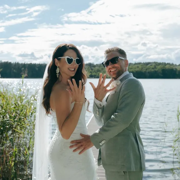 Brautpaar mit Sonnenbrillen zeigt stolz die Eheringe am Seeufer.