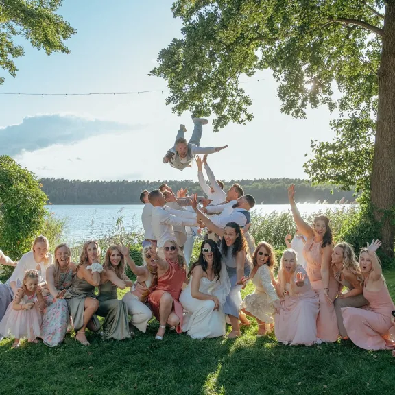Gruppe fröhlicher Menschen in festlicher Kleidung posiert im Grünen am See, Baum im Hintergrund.
