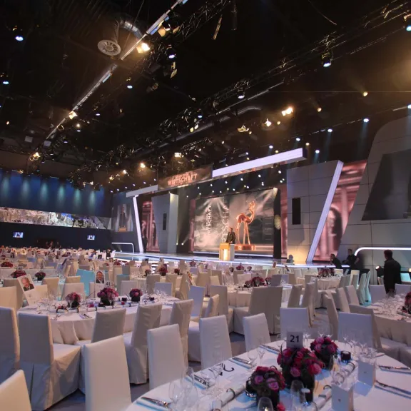 Großer, elegant dekorierter Veranstaltungssaal mit vielen runden Tischen und lila Blumen.