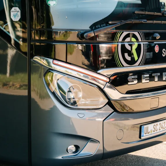 (2)	Nahaufnahme der Front eines modernen grauen Reisebusses von „Schröder Reisen“ mit Setra-Logo, Scheinwerfern und Kennzeichen.
